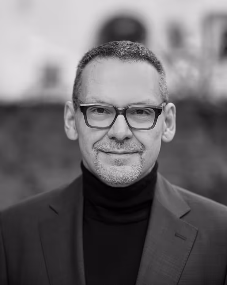 Smiling man wearing glasses, a turtleneck, and a blazer, with a blurred outdoor background.