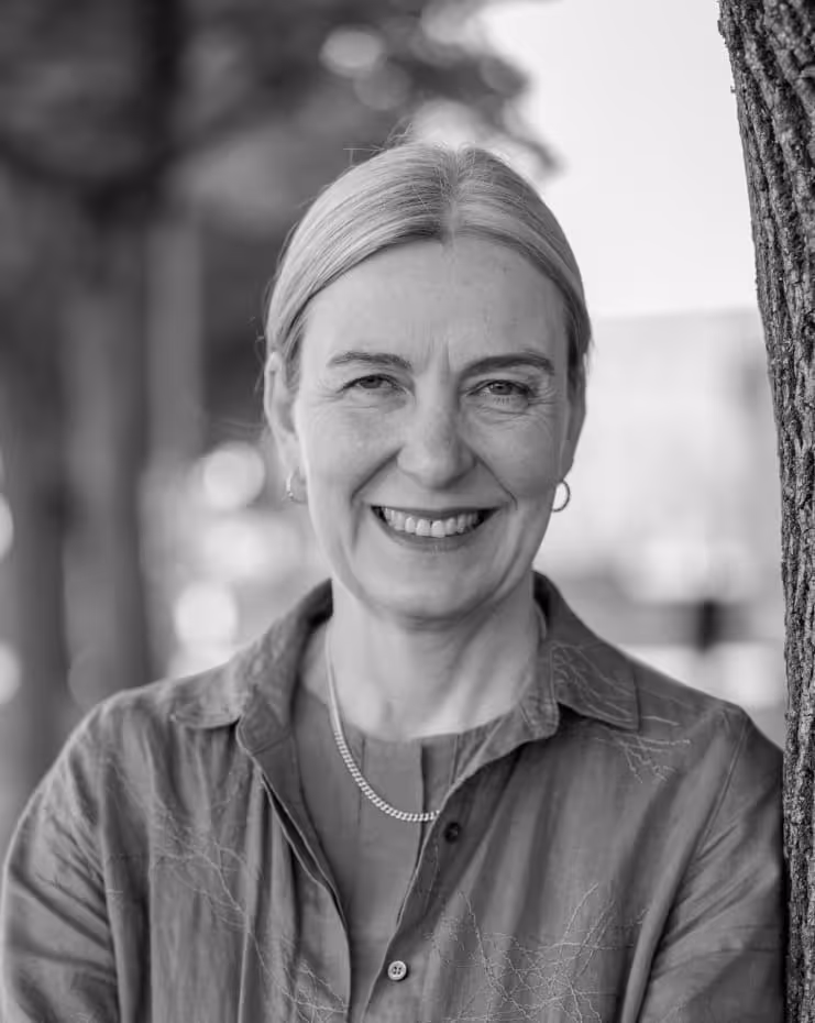 Smiling woman with light hair, wearing a buttoned shirt and necklace, standing outdoors near a tree.