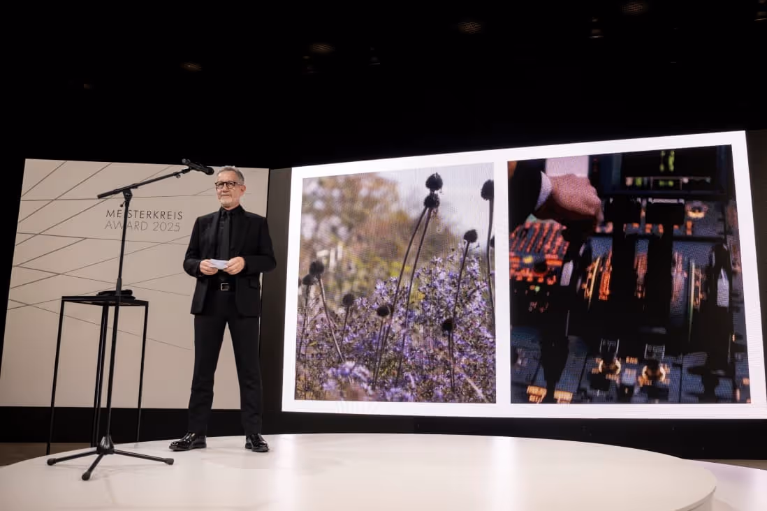 Man in black suit standing on stage at a microphone with a backdrop displaying 'MESTERKREIS AWARD 2025' and two large images, one of flowers and the other of a hand on airplane controls.
